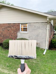 a hand holding a paint brush in front of a brick house with grass and bushes