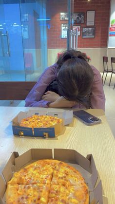 two pizzas in boxes on a table with a woman sleeping next to them and one has her head down