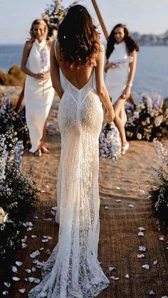 three women in white dresses walking down a path near the ocean with flowers all around them