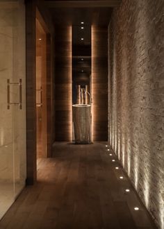 a long hallway with stone walls and lights on either side of the wall, leading to another room