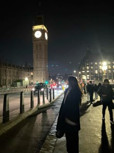 a woman standing on the sidewalk in front of a clock tower at night with people walking by