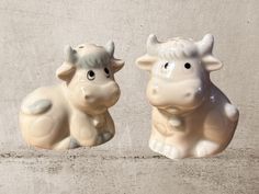 two ceramic cows sitting next to each other on a cement surface with concrete wall behind them