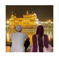 two people sitting on the edge of a body of water looking at a golden building