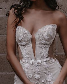 a woman wearing a wedding dress with flowers on the skirt and in front of a brick wall