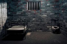 a prison cell with a bed and jail bars on the wall, in front of a brick wall