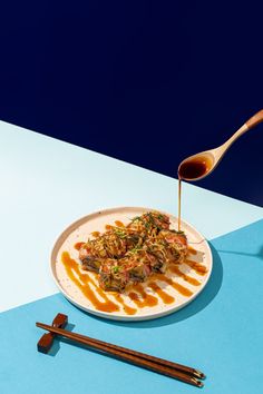 a white plate topped with meat and sauce next to chopsticks on a blue table