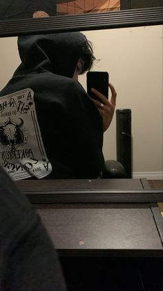 a person wearing a hoodie looking at their cell phone in front of a mirror