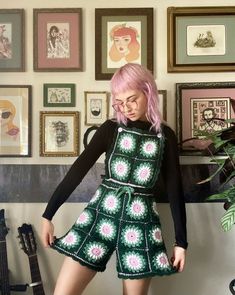 a woman with pink hair wearing a green crochet dress in front of a wall full of framed pictures