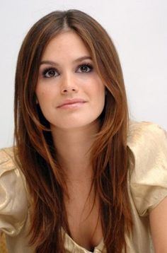 a woman with long brown hair sitting in front of a white wall and looking at the camera