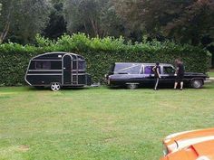 two people standing in front of a black trailer and an orange car on the grass