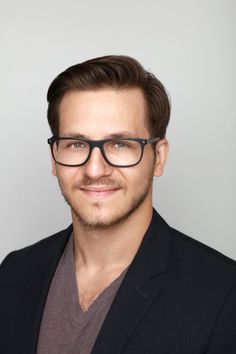 a man with glasses and a suit jacket is smiling for the camera while standing in front of a gray background