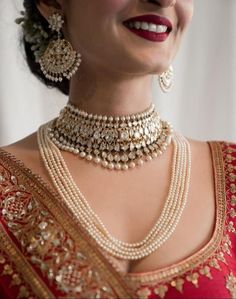 a woman in a red and gold sari with pearls on her neck, smiling at the camera