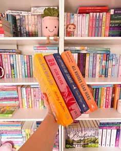 a person holding up three books in front of a book shelf filled with many different colored books