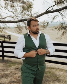 a man standing in front of a fence wearing a green suit and white shirt with his hands on his hips