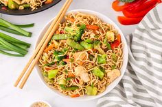 a bowl filled with noodles and vegetables next to chopsticks on a white table