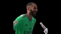 a man with a beard holding a microphone and wearing a green soccer jersey, smiling at the camera