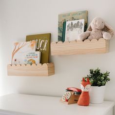 two wooden shelves holding books and toys on top of a white shelf next to a potted plant