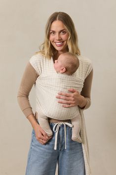 a woman is holding a baby in a white sling and smiling at the camera while wearing jeans