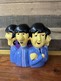 three ceramic figurines are sitting on a wooden table, one has his arms around the other's neck