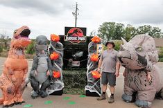 two men standing next to three inflatable dinosaur costumes on the street with trees and bushes behind them