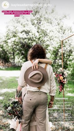 two people standing next to each other in front of trees and grass with flowers on the ground