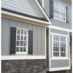 a gray house with two windows and shutters on each side, along with a stone wall