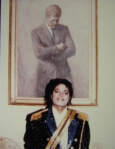 a man in a suit and tie standing next to a portrait of elvis presley with his arms crossed