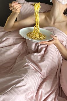 a woman laying in bed while eating spaghetti from a plate with a fork and knife