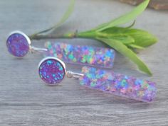 two purple and blue earrings sitting on top of a wooden table next to green leaves