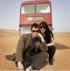 two people sitting on the ground in front of a red double - decker bus,