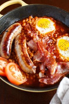 eggs, beans and tomatoes are in a skillet with bacon on the side next to tomato slices