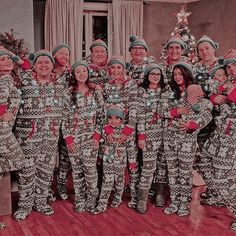 a group of people standing next to each other wearing matching pajamas and hats in front of a christmas tree