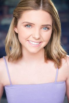 a young woman with blonde hair and blue eyes smiles at the camera while wearing a purple tank top