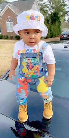 a baby wearing overalls and a hat standing on the hood of a black car