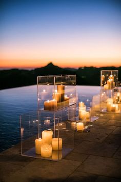 candles are lined up along the edge of a pool at dusk, with water in the background