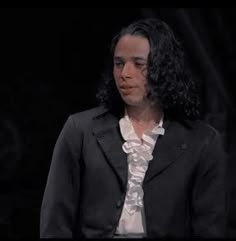 a man with long hair wearing a black suit and white shirt is standing in front of a dark background