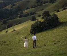 a man and woman walking across a lush green field next to a tree filled hillside