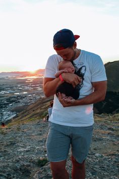 a man holding a baby while standing on top of a hill with the sun setting in the background