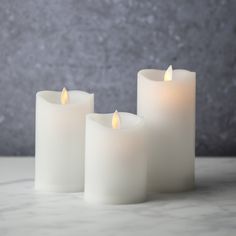 three white candles sitting next to each other on a marble counter top with a gray wall in the background