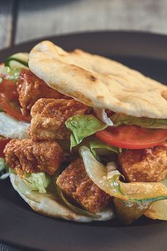 a chicken sandwich with lettuce, tomato and onions on a black plate next to pita bread