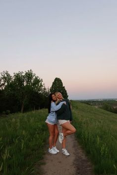 two young women hugging each other on a dirt path in the middle of a field