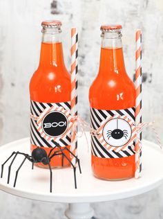 two orange soda bottles sitting on top of a white cake plate with black spider decorations