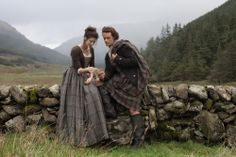 two people standing next to each other in front of a stone wall with mountains behind them