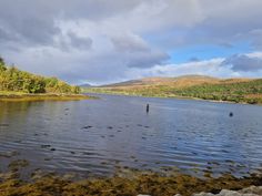 a body of water surrounded by trees and mountains under a cloudy blue sky with clouds