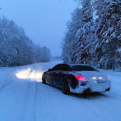 a car that is sitting in the snow near some trees and bushes, with it's lights on