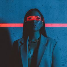 a woman with red eyes wearing a black suit and chain necklace standing in front of a blue wall