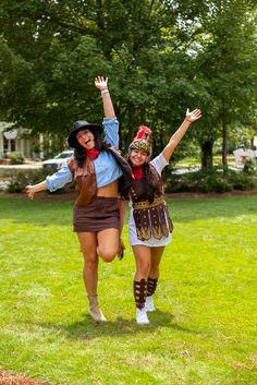 two women dressed in costumes are walking through the grass with their arms up and hands raised