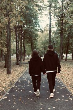 two people walking down a path in the middle of a park with leaves on the ground