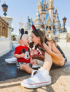 a woman and her baby are sitting in front of the castle at disneyland world with mickey mouse