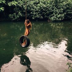 a woman hanging from a rope while riding a tire in the water with trees behind her